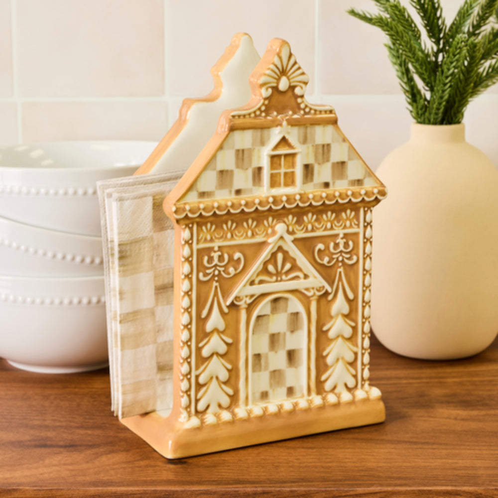 A gingerbread house-shaped napkin holder with a warm beige and brown checkered pattern, standing on a wooden countertop with kitchen bowls and a vase in the background.