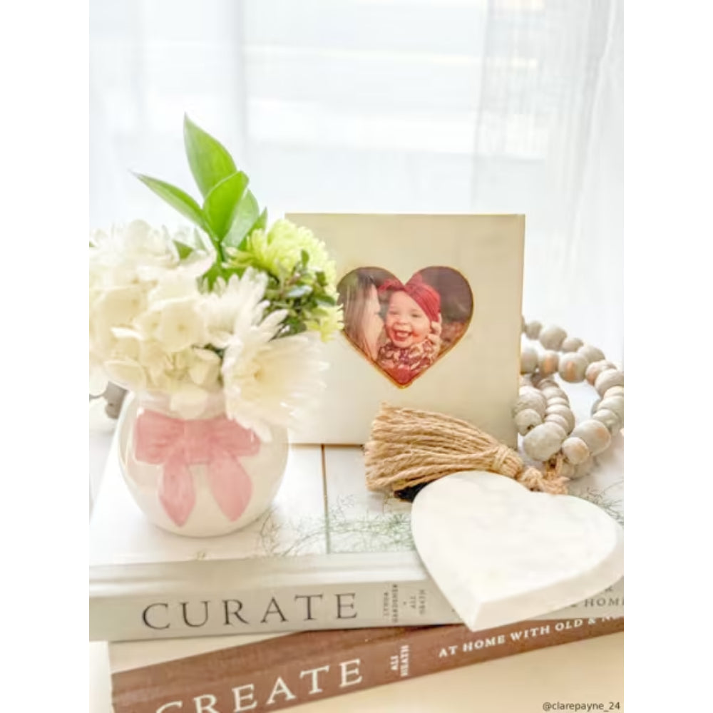 A styled tabletop arrangement featuring a cream-colored photo card with a heart-shaped cutout holding a child's portrait, placed on top of stacked books beside white flowers in a pink vase, a tassel, a wooden bead garland, and a white heart-shaped object.