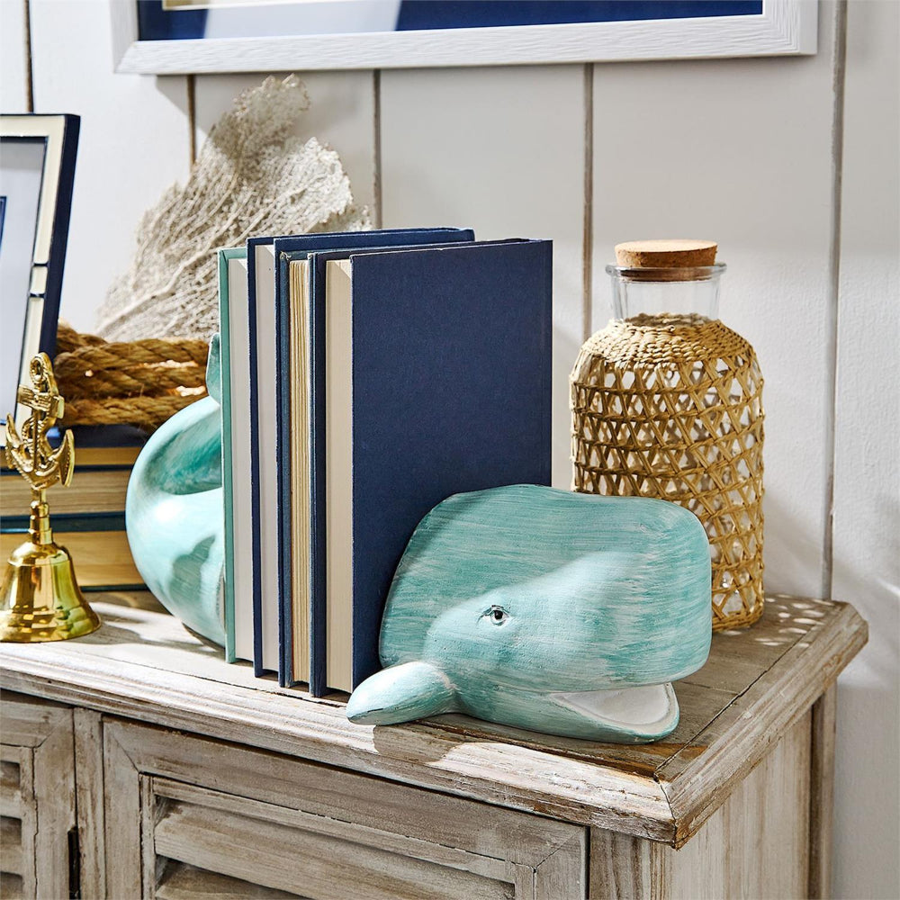 A aqua whale-shaped bookend holding a stack of navy and beige books on a rustic wooden console table, styled with a woven glass bottle, a gold bell, and a picture frame against a white paneled wall.