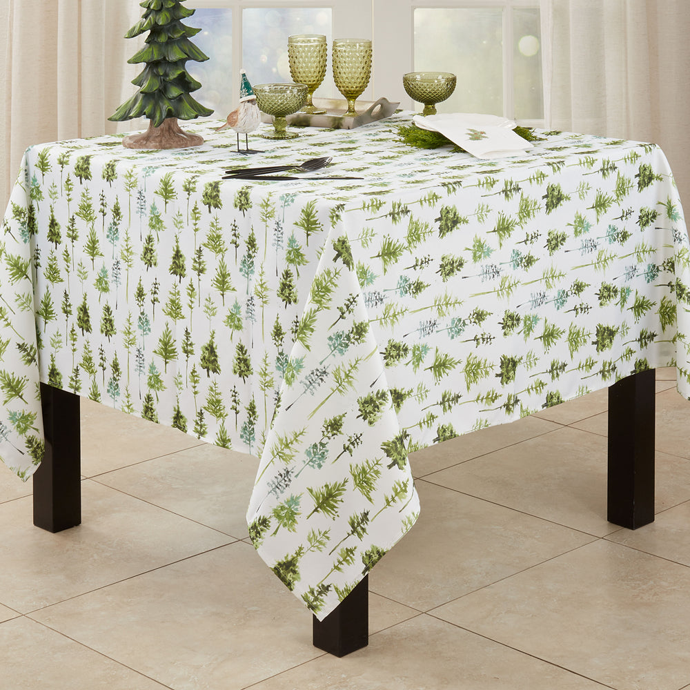 A rectangular table covered with a white tablecloth printed with a repeating pattern of green pine trees and small botanical motifs. The table is styled with green textured goblets, a small decorative tree, and other tableware in a bright dining room setting.