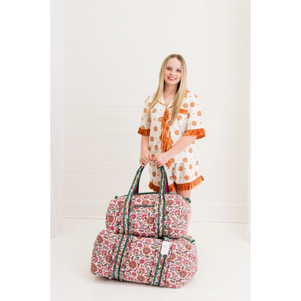 A woman wearing a cream pajama set with orange ruffles stands in a bright room holding two stacked floral duffel bags with green striped handles.