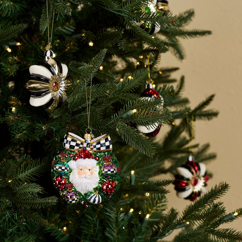 A Christmas ornament shaped like a green wreath with a white Santa face in the center, red and striped ornaments, and a black-and-white checkered bow at the top on a christmas tree.