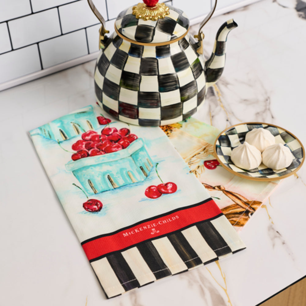 A kitchen towel with a black and white checkered border and a red band reading 'MICKERENZ CHILDS' lies on a marble countertop next to a matching checkered teapot and a small plate of meringues.