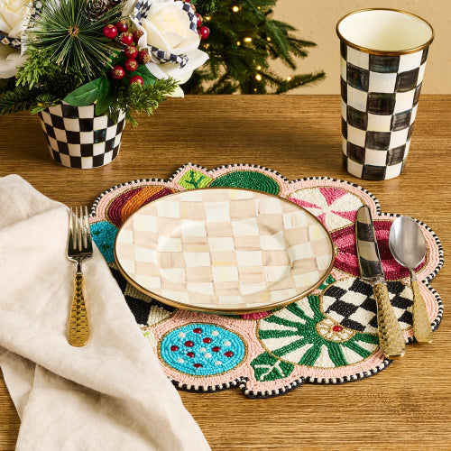 A styled tabletop setting with a decorative multicolor placemat, a cream checkered oval plate, matching checkered napkin and cup, gold-toned cutlery, and holiday greenery in the background.