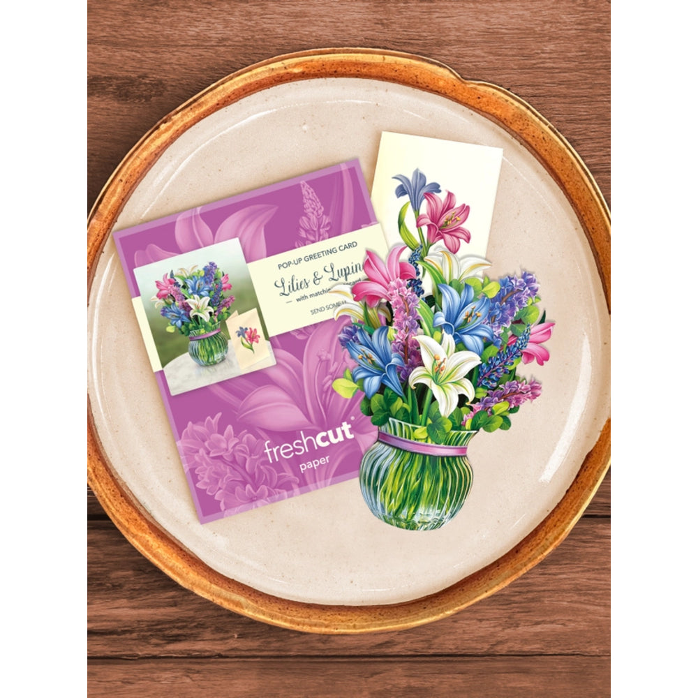 A flat-lay product shot of a 'fresh cut' pop-up greeting card set on a round ceramic plate with a gold rim, set against a dark wood background. The set includes a purple card with floral graphics and a small illustrated card showing a bouquet of pink, blue, and white flowers in a green vase.