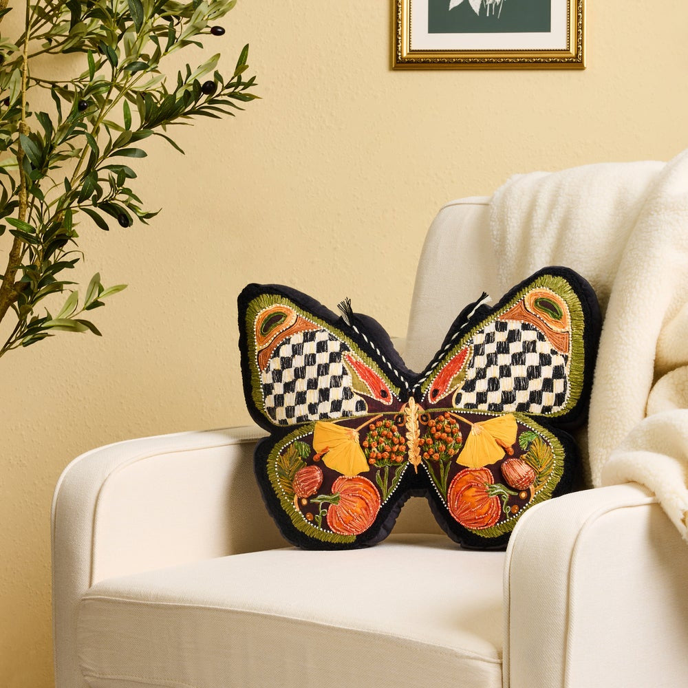 A decorative butterfly-shaped throw pillow with embroidered autumn motifs and checkered wings, resting on a cream armchair in a styled room setting.