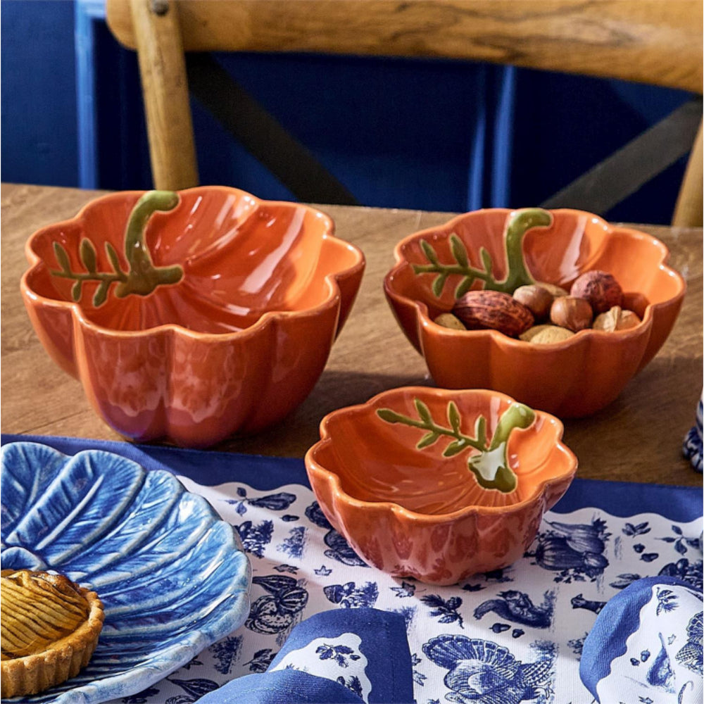 Three pumpkin orange ceramic bowls with scalloped edge with green leaf accents arranged on a wooden table, with one bowl containing nuts. A blue patterned cloth and a blue plate are partially visible in the foreground.