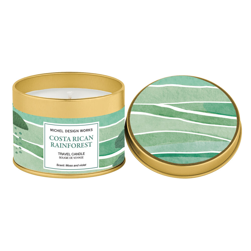 A travel candle in a round gold metal tin with a matching lid placed beside it on a white background. The tin and lid feature a watercolor-style green landscape design with the label reading 'MICHEL DESIGN WORKS COSTA RICAN RAINFOREST'.