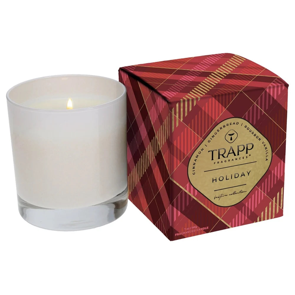 A lit white glass candle next to its red and gold plaid packaging box, featuring a gold TRAPP Fragrances Holiday label.