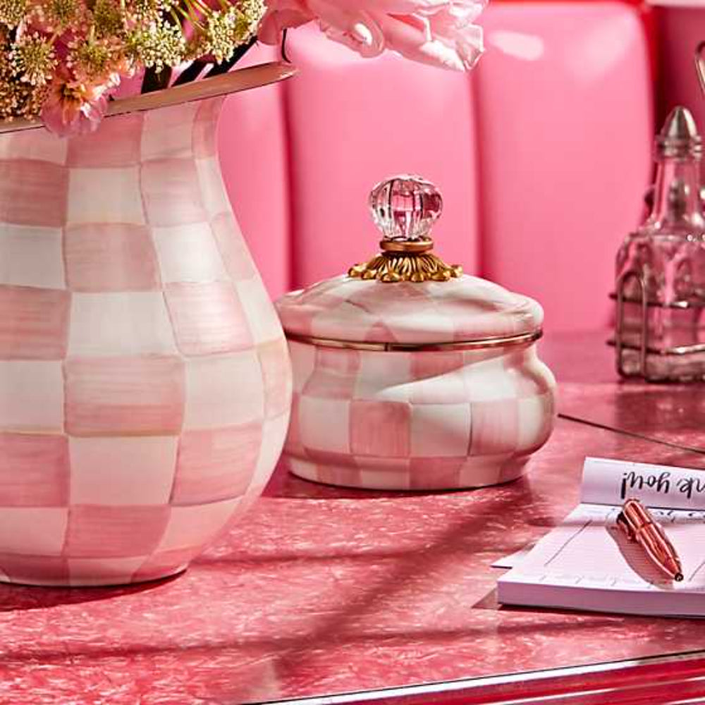 Life style photo of a decorative jar with a rosy pink and white checkered pattern and gold accents on a table with a pink backround with others.