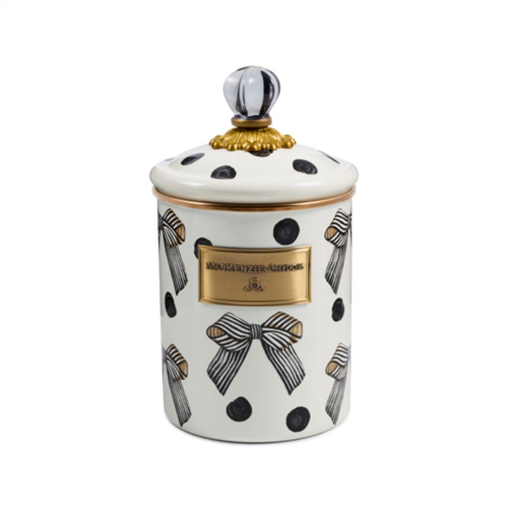 A white enamel canister with a matching lid, decorated with black polka dots, striped bows, and gold accents, including a gold brand plaque on the front.