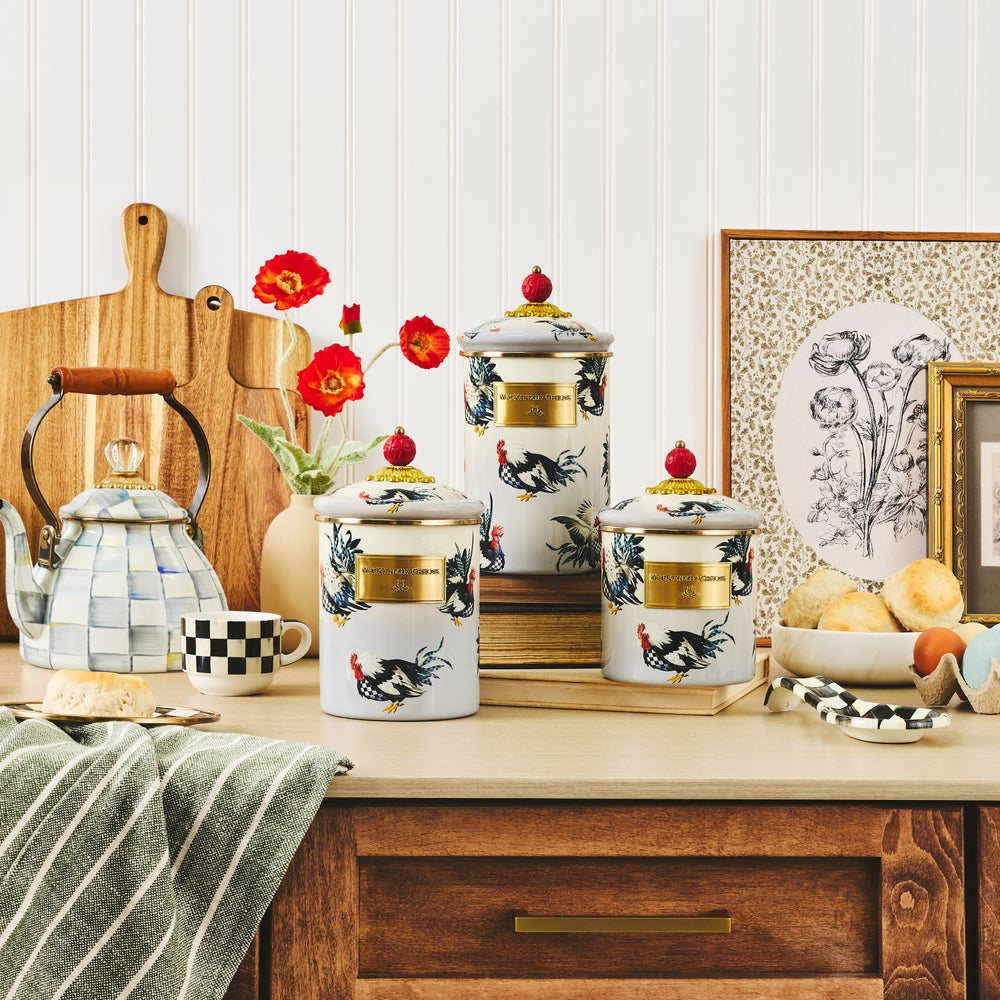 Three white canisters with blue hen designs and a red top on a tan table with other kitchen items against a white wall.