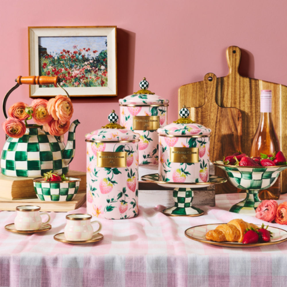 A styled tea-time scene with pink and white patterned canisters, a green-and-white checkered teapot, cups, bowls of strawberries, croissants, flowers, and a pink wall background.