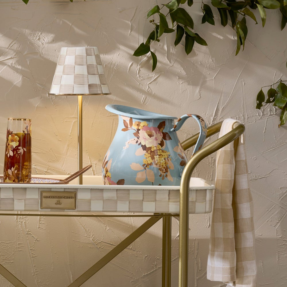 A light blue floral pitcher with a gold handle, displayed on a gold-framed tray table in a styled interior setting with a checkered lamp, a checkered towel, and a textured beige wall.