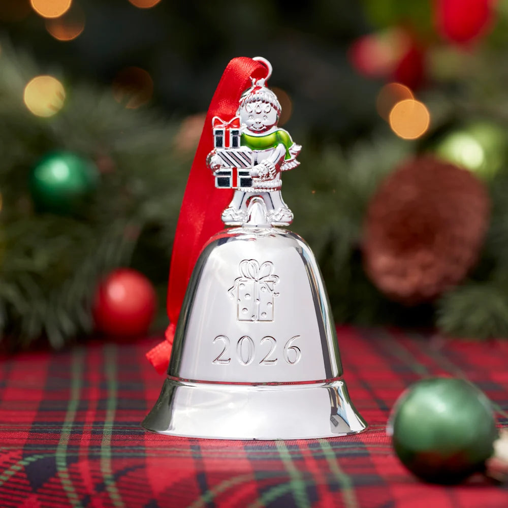 Silver bell ornament with Santa figure and '2026' on a plaid surface with Christmas decorations in the background.