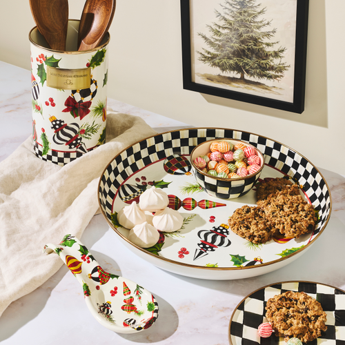 A styled tabletop scene with a large oval holiday-themed serving platter, a matching small plate, a spoon, and a utensil holder. The platter and holder have a white base with black-and-white checkered trim, red berries, green holly, plaid accents, and festive illustrations.