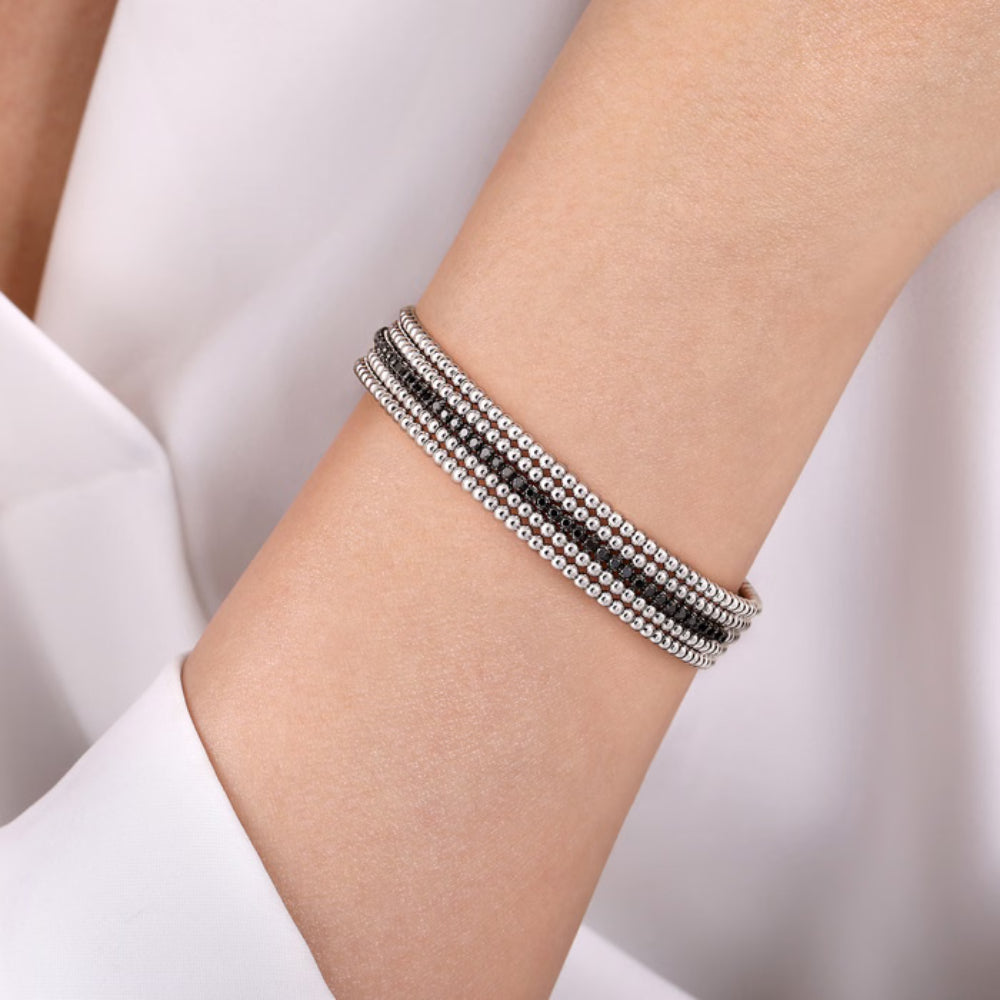 A close-up of a person's wrist wearing a multi-strand bracelet with silver-toned and black stones, set against a soft white background.