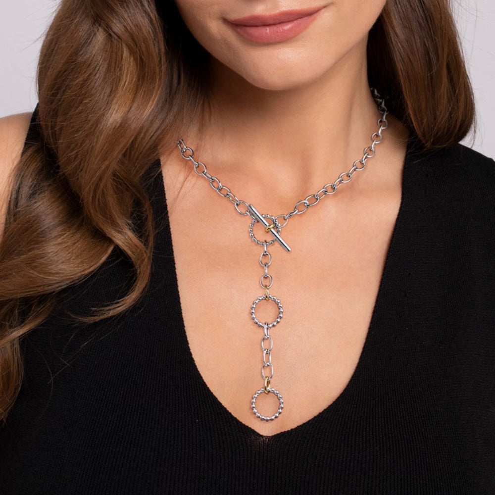 A close-up of a woman wearing a silver-toned lariat-style necklace with a toggle clasp and three dangling circular links, paired with a black top.