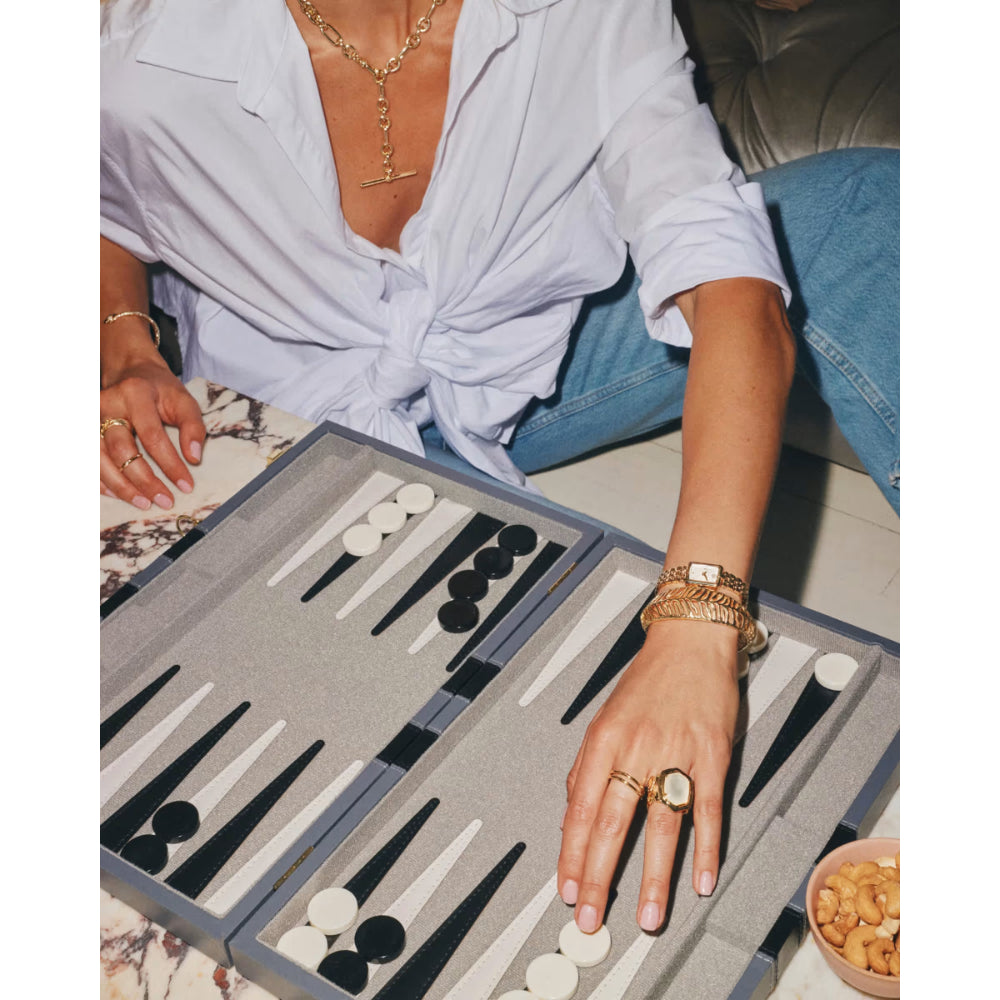 A close-up lifestyle shot of a person playing a game of backgammon, wearing a white shirt and blue jeans, with a gray backgammon board and black and white checkers in the foreground.