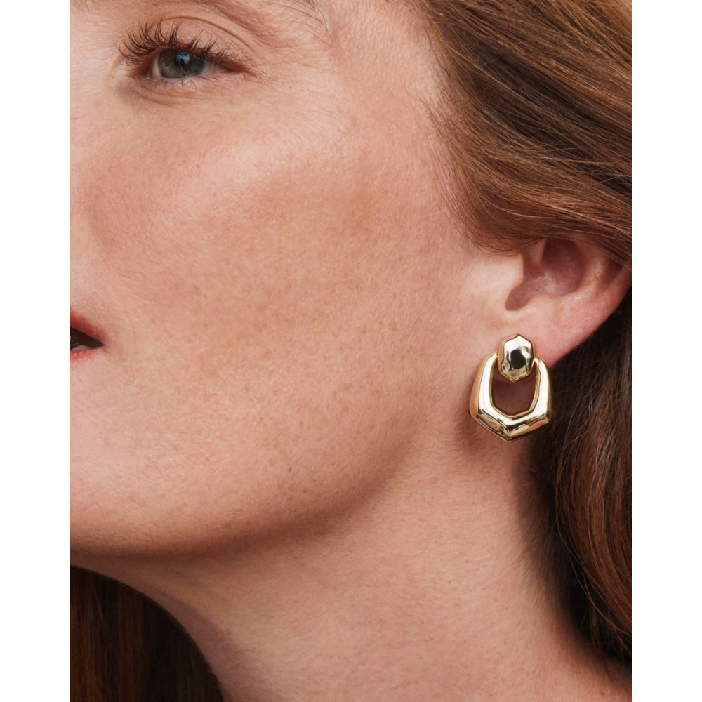 A close-up of a model's ear wearing a gold geometric hoop earring with a small faceted green gemstone at the top.
