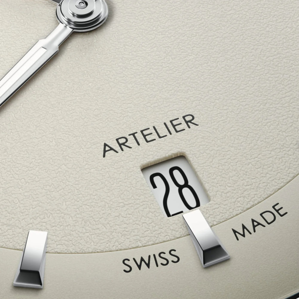 Close-up of a watch dial with a textured light beige face, silver metal hands and markers, and a window showing the number 28. The words ARTELIER, SWISS, and MADE are printed on the dial.