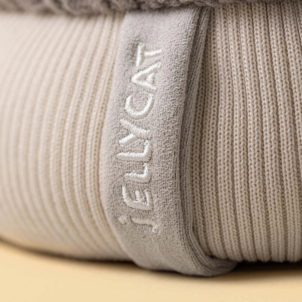 Close-up of a ribbed knit hat in a light warm gray-beige color, with the brand name 'JELLYCAT' embroidered vertically on a fabric tab.
