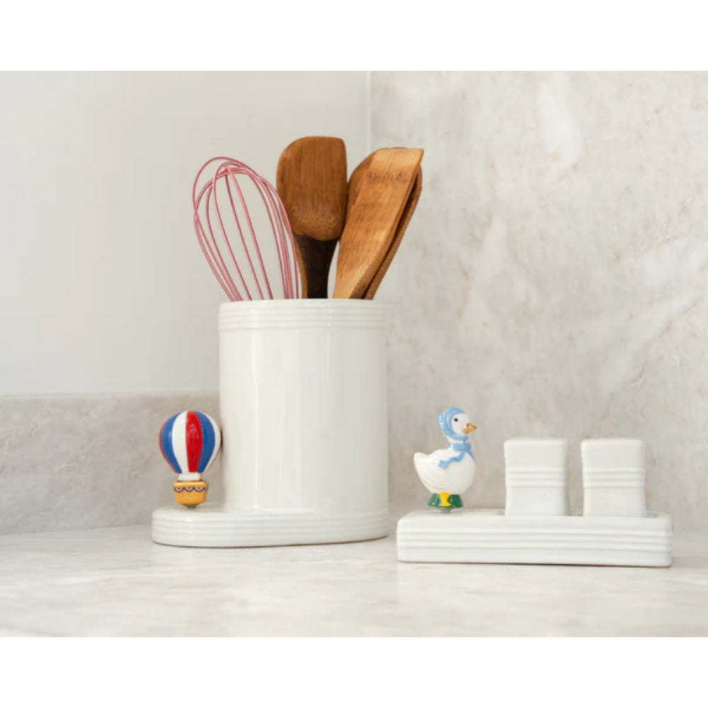 White kitchen utensil holder with wooden spoons, whisk, and small decorative items on a marble countertop.
