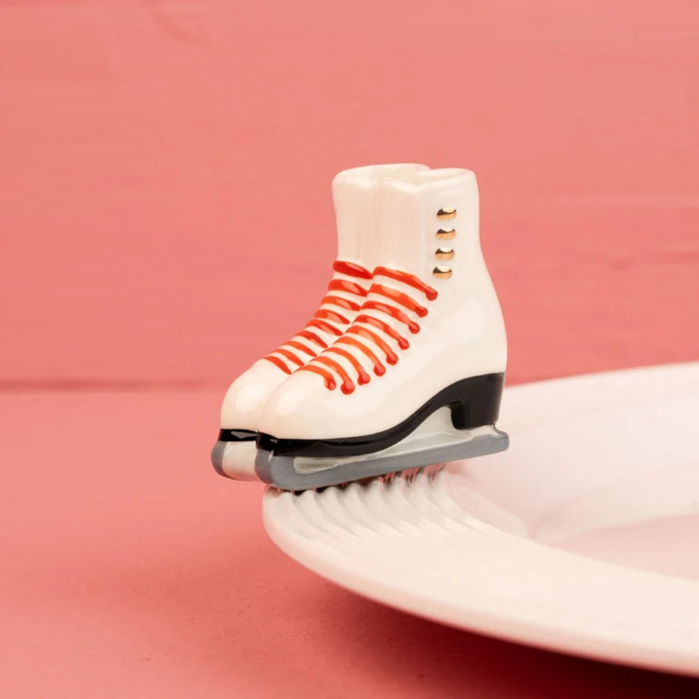 A miniature white ice skate figurine with red laces and a black blade, balanced on the edge of a white plate against a pink background.
