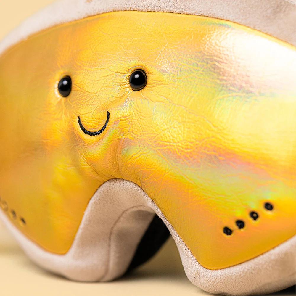 A close-up of a plush ski goggles or face shield made from iridescent yellow and gold leather, featuring a stitched smiling face and black eye details, with beige fabric trim.