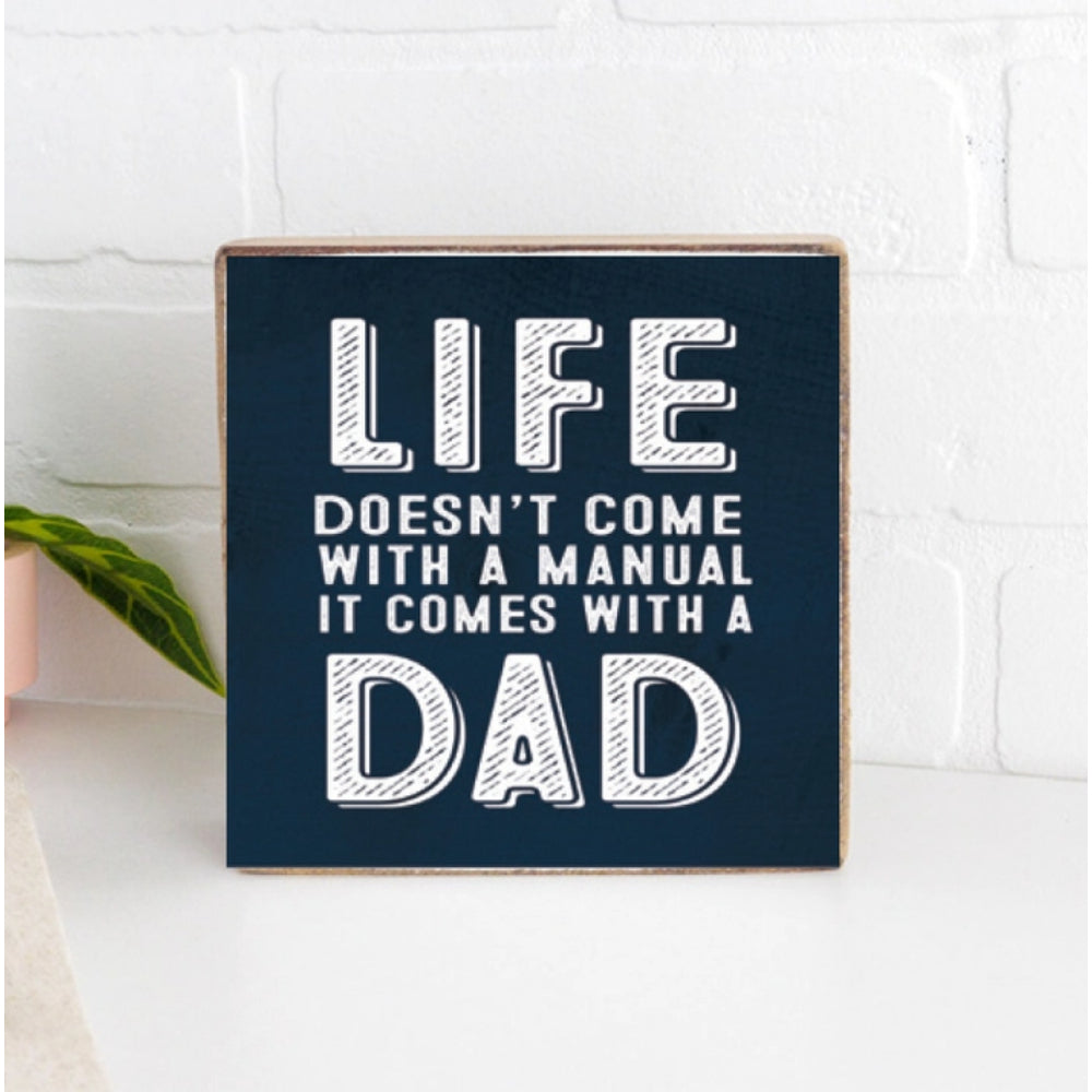 Navy blue wood square decorative block sign with motivational quote in white text that reads " Life Doesn't come With A Manual It Comes With A Dad" on a kitchen counter with a white background