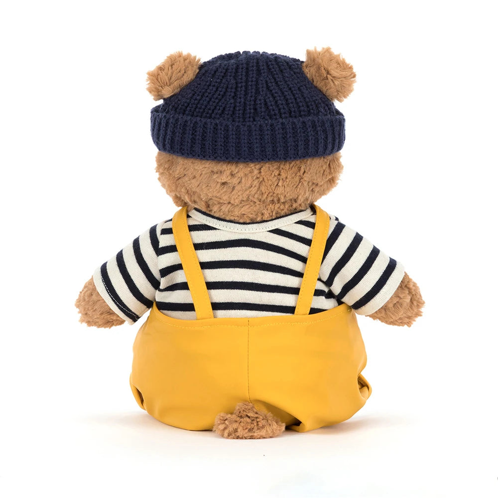 A photo of the back of a beige teddy bear with a colorful seafarer outfit with yellow overalls,navy cap and a white and blue striped shirt on a white backround