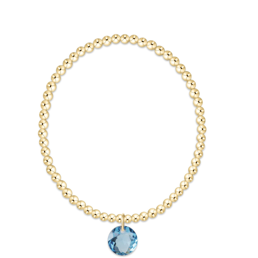 A gold beaded bracelet with a single faceted light blue gemstone pendant, displayed on a clean white background.
