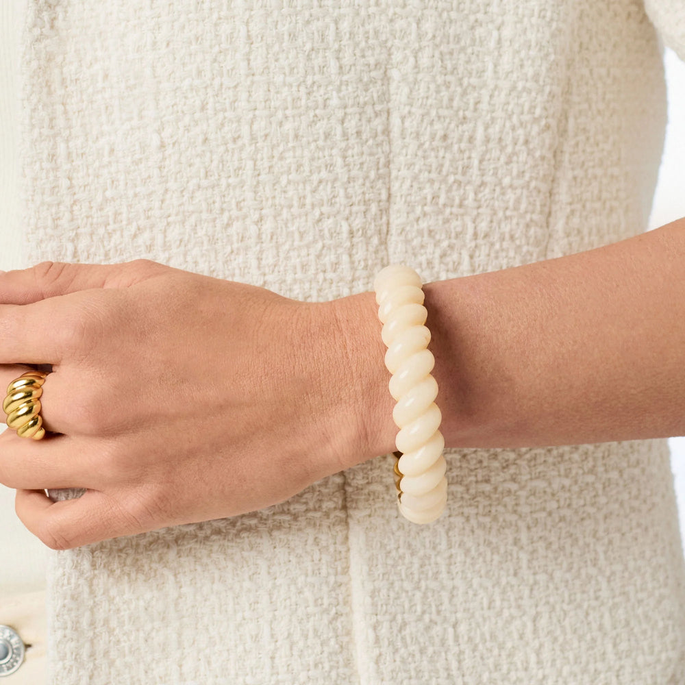 A close-up of a person's wrist wearing a thick, twisted cream-colored bangle bracelet, paired with a matching textured cream jacket and a gold ring.