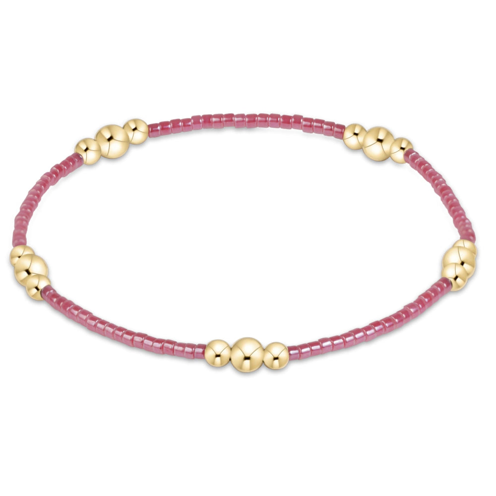 A delicate beaded bracelet with a pinkish-red cord and small gold metallic beads, shown against a plain white background.