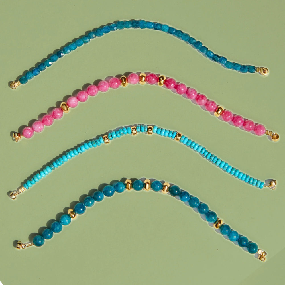 Four beaded stretch bracelets arranged in curved rows on a light olive-green background. The bracelets feature faceted beads in shades of blue, pink, and turquoise, separated by small gold spacer beads, and finished with gold-toned clasps.