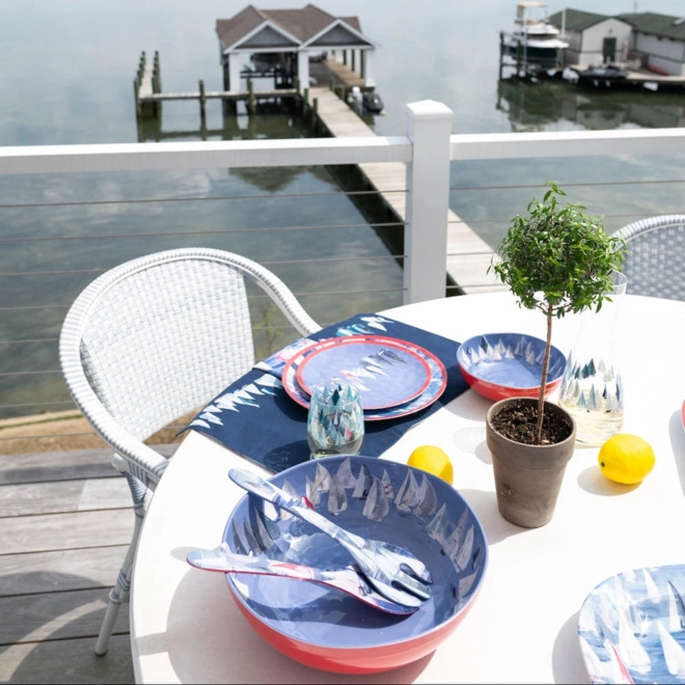 Outdoor dining table set with blue and red plates, silverware, and a lemon on a deck by a waterfront.