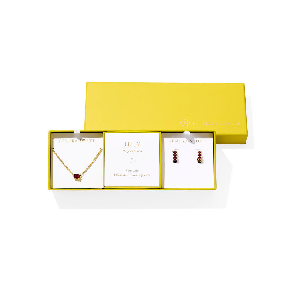 A Kendra Scott jewelry gift set in a bright yellow box, featuring a gold necklace with a burgundy crystal pendant, matching burgundy-and-gold earrings, and a central card reading 'JULY'.