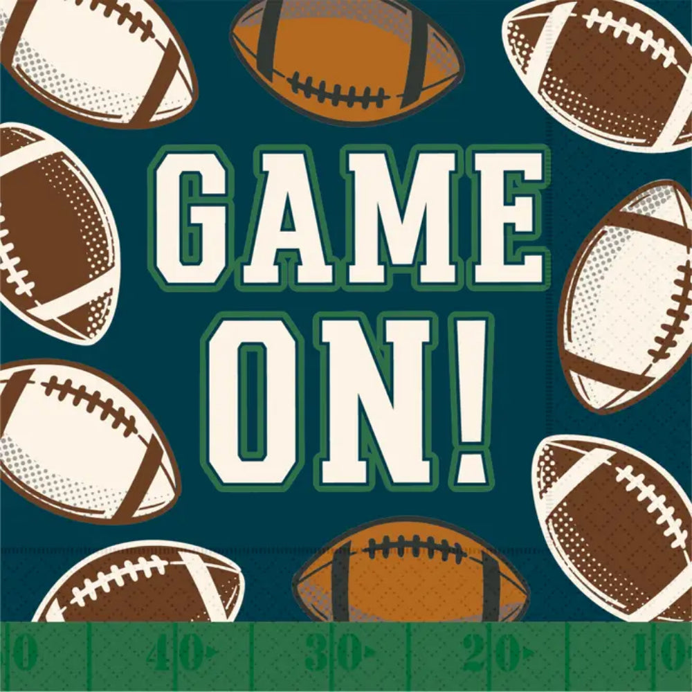A square football-themed napkin with the words “GAME ON!” in large white block letters outlined in green, centered on a dark teal background. Brown and cream footballs are scattered around the edges, and a green yardline strip appears along the bottom edge.