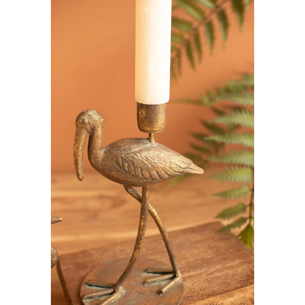 A bronze-toned metal candlestick holder shaped like a walking bird, holding a white taper candle, set on a wooden surface against a warm orange-brown background with blurred green leaves.