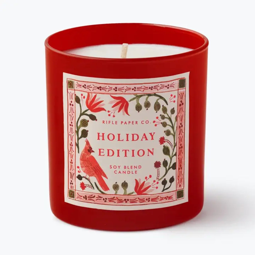 A red glass jar candle with a white wick and a decorative front label reading 'Rifle Paper Co. Holiday Edition Soy Blend Candle,' featuring a red cardinal and floral illustration on a white background.