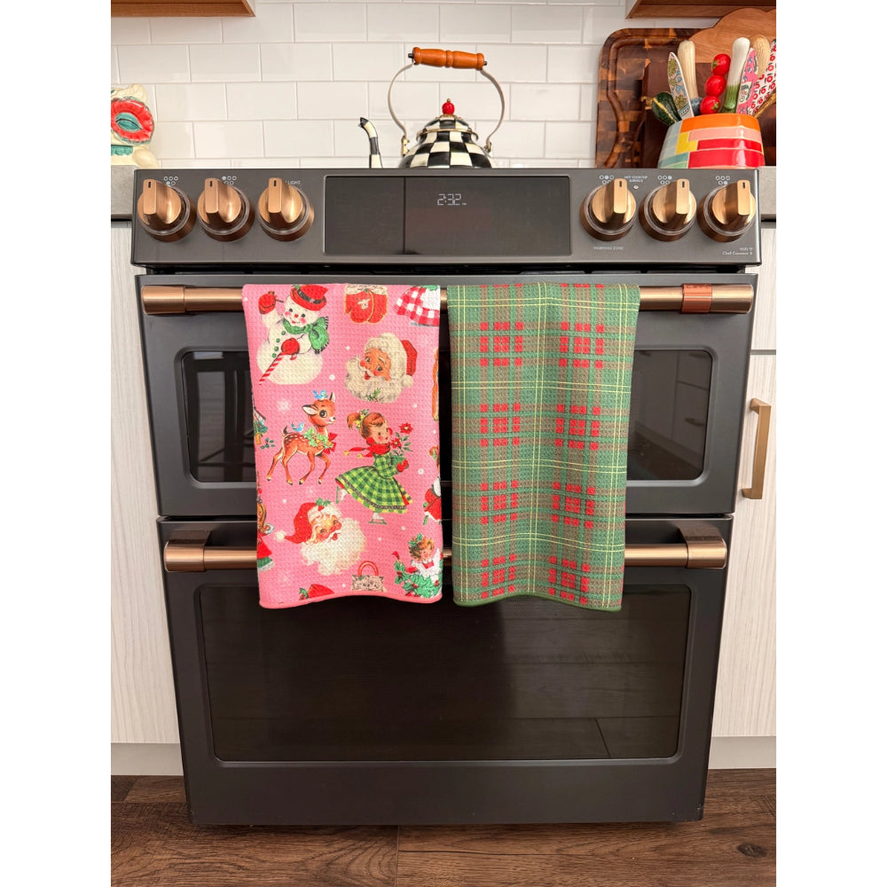 Two holiday-themed kitchen towels hanging on the handles of a black double oven stove. The left towel is pink with Santa Claus and reindeer illustrations, and the right towel is green with a red plaid pattern.