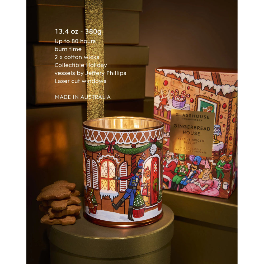 A holiday-themed candle vessel with a gingerbread house illustration, shown next to its branded packaging box and a small stack of gingerbread cookies on a dark shelf.