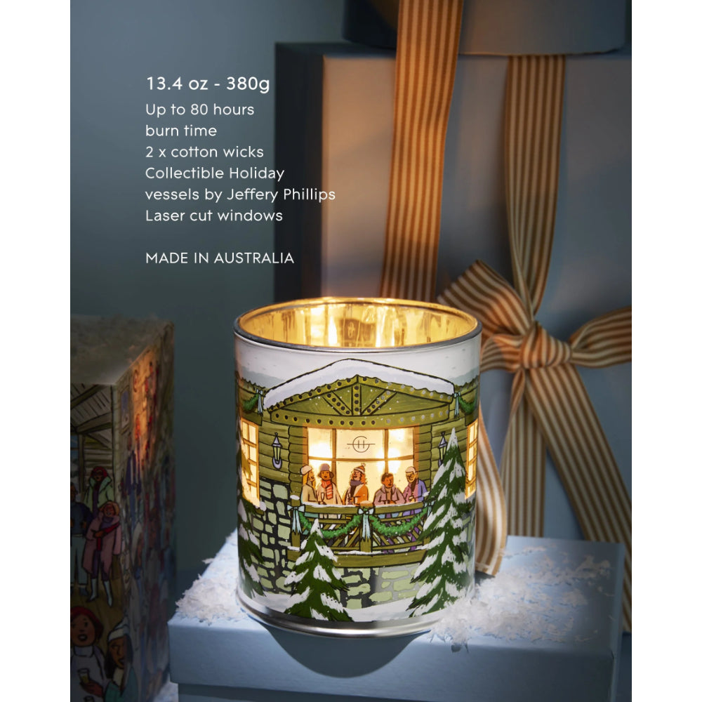 A holiday-themed collectible candle in a cylindrical glass vessel featuring a painted Christmas house scene with trees and warm glowing windows. The candle is displayed on a blue pedestal with gift boxes and ribbon in the background, with product text visible on the left.