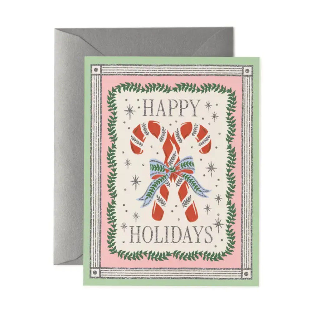 A square holiday greeting card with a gray envelope behind it, featuring the words “HAPPY HOLIDAYS” around an illustration of two red-and-white candy canes with green and blue accents on a pale pink background.