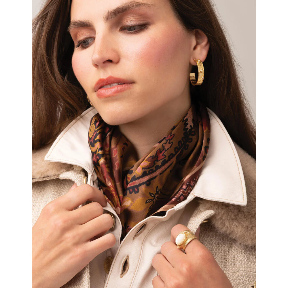 A close-up lifestyle shot of a woman wearing a patterned silk scarf tied around her neck, a cream-colored coat with a fur collar, and gold jewelry including a hoop earring and ring.