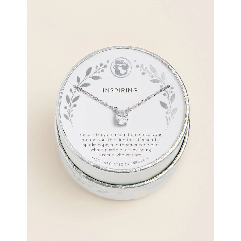 A silver-plated necklace with a clear crystal pendant is displayed on the inside of a round silver gift box. The white insert card features the word 'INSPIRING' and a silver leaf border.
