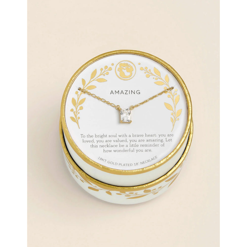 A gold-plated 18k necklace with a clear gemstone pendant, displayed on a white card inside a round white and gold presentation box. The card features the brand name 'AMAZING' and a sentimental message, set against a warm cream background.