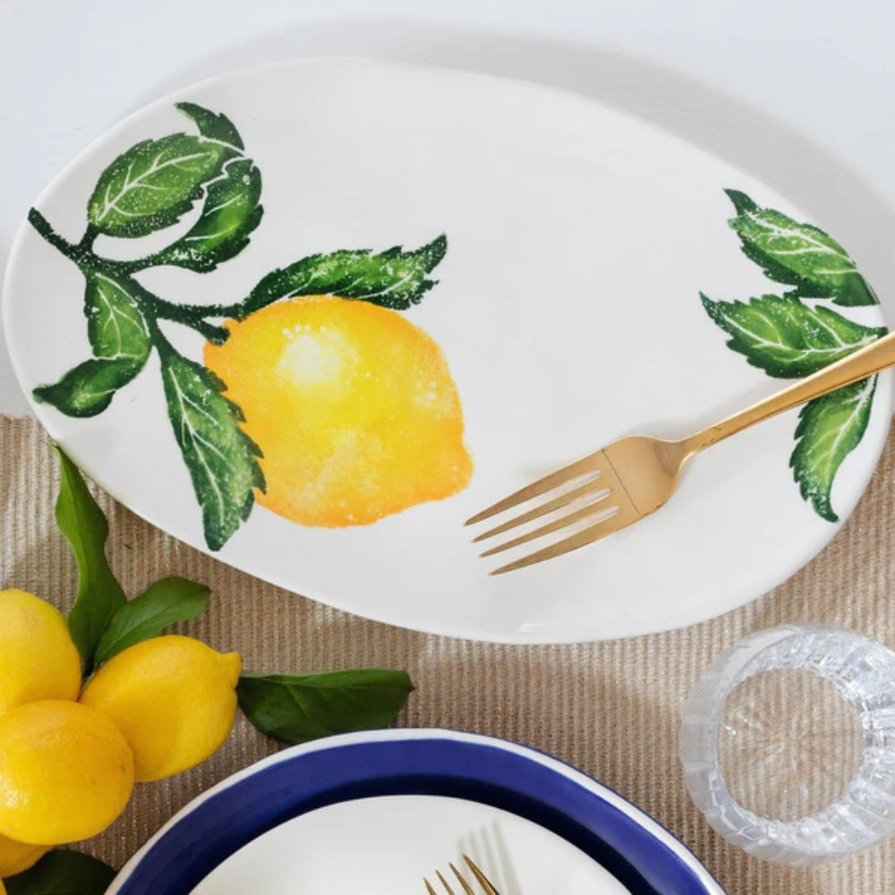 A small oval platter featuring a handpainted lemon and green leaves design on a tan tablecloth table with a gold folk on it with lemons and glass as well as a plate and with a blue rim.