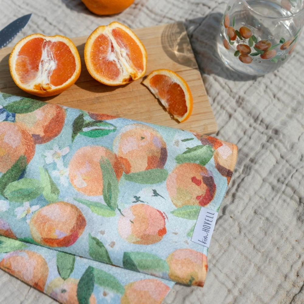 Blue tea towel with orange pattern on a wooden cutting board with cut oranges.