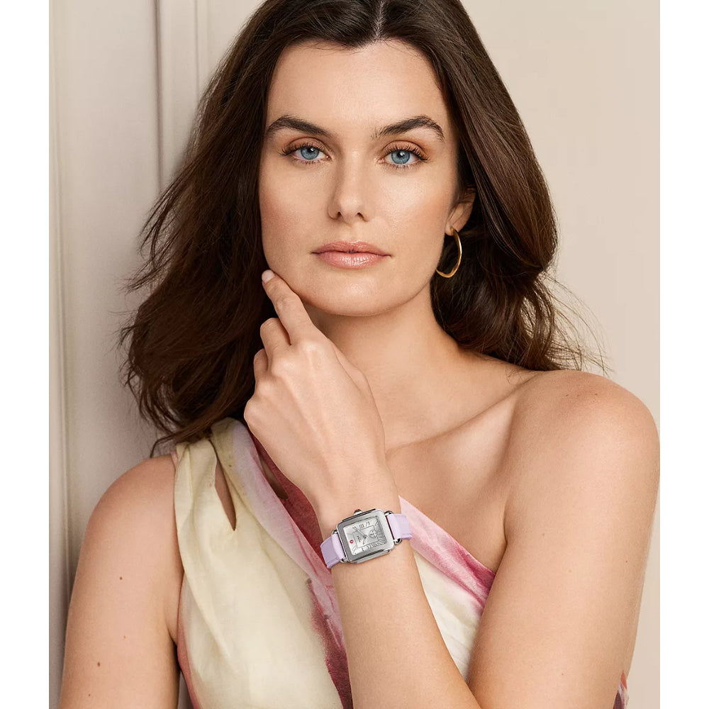 A woman with wavy brown hair poses in a cream and pink draped top, wearing a silver-toned rectangular watch with a light pink strap. She is set against a soft beige background.